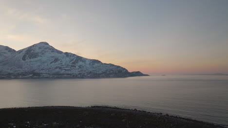 Eine-Langsame-Drohnenaufnahme,-Die-über-Einem-Ufer-Der-Insel-Kvaloya-In-Norwegen-Fliegt,-Mit-Schneebedeckten-Berggipfeln-Im-Hintergrund-Und-Einem-Rosafarbenen-Sonnenuntergang-Am-Horizont