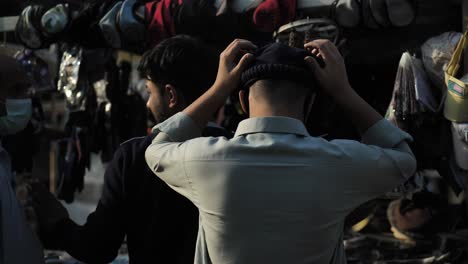 Back-View-Of-Male-Trying-On-Hat-Cap-At-Market-In-Karachi,-Pakistan