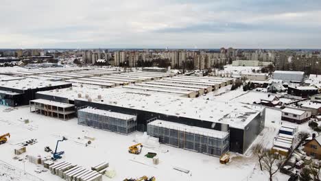 Logistics-building-under-construction-with-Kaunas-city-residential-district-behind-during-snowfall,-aerial-ascend-view