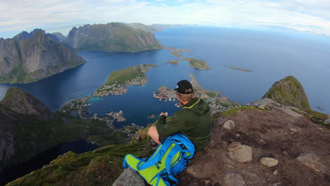 Traveler-man-sitting-on-a-hill-of-beautiful-norway-fjord-Reinebringen-near-Reine-town,-Lofoten-Islands