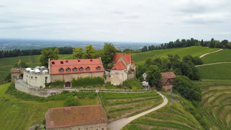 Aerial-orbit-of-chateau-on-top-of-a-green-hill-and-wine-vineyard-terraces-surrounded-by-forest,-mountains-in-background-on-an-overcast-day,-Switzerland