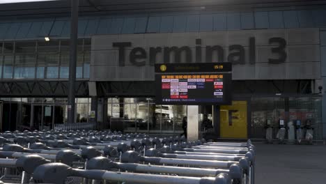 Reihe-Von-Gepäckwagen-Vor-Dem-Heathrow-Terminal-3-Gebäude-Mit-Abflugtafel-Der-Fluggesellschaft