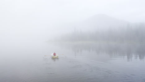 Person-rowing-a-boat-in-white,-foggy-lake-landscape-with-mountain-and-forest-in-the-background