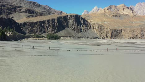 aerial-drone-flying-backwards-revealing-the-Passu-Cones-mountains-in-the-distance-as-tourists-cautiously-cross-the-famous-Hussaini-Bridge-in-Hunza-Pakistan-as-a-fast-paced-gray-river-flows-below