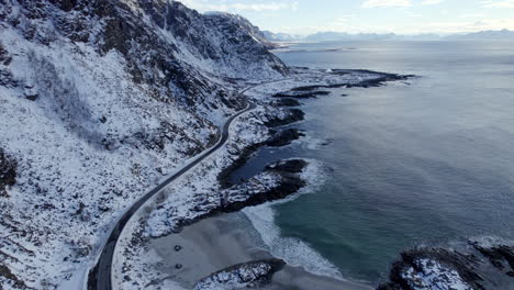 Aerial-dolly-shot-along-the-Scenic-Route-Andøya-with-waves-crashing-on-the-beach-and-snow-covered-mountains