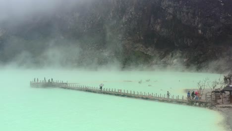 aerial-drone-of-a-green-toxic-sulfur-lake-inside-a-volcano-called-Kawah-Putih-with-fumes-and-gasses-rising-while-tourists-walk-a-long-bridge-in-Bandung-Indonesia
