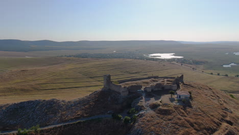 Long-drone-tracking-shot-of-Enisala,-medieval-fortress-on-top-of-a-hill-surrounded-by-lakes-and-plains-on-a-clear-sunny-day