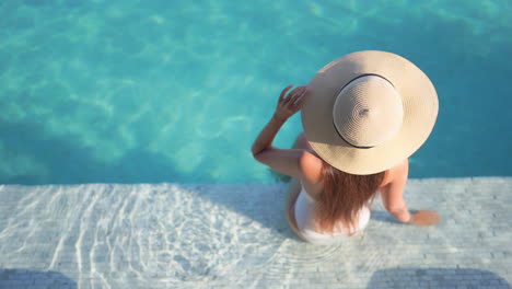 Back-of-Sexy-Woman-in-Swimsuit-and-Floppy-Summer-Hat-by-Swimming-Pool-on-Sunny-Tropical-Day