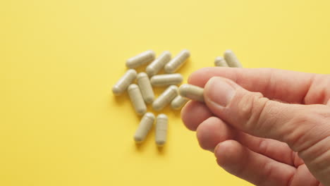 Right-Male-Hand-Holding-Lions-Mane-Supplement-Pill-In-Between-Fingers-With-Yellow-Background-With-Pills-On-Table