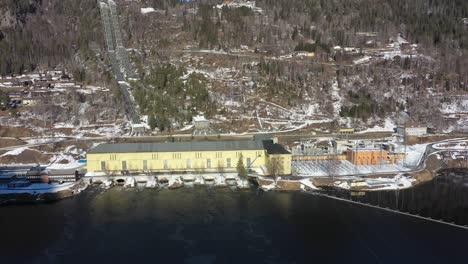 Nore-I-hydroelectric-powerplant-operated-by-Statkraft-in-Rodberg-Norway---Sideways-moving-sunny-day-aerial-overview-with-pipelines-coming-down-hillside-in-background