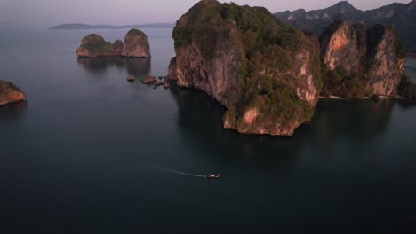 Wide-high-altitude-aerial-drone-of-unique-limestone-mountain-formations-near-Railay-Beach-in-Krabi-Thailand-during-sunrise-as-a-thai-longtail-boat-motors-in-the-Andaman-Sea