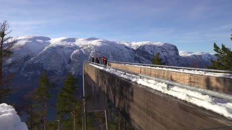 Tourists-enjoying-stunning-view-on-edge-of-Stegastein-viewpoint-Norway---Sideways-moving-clip-behind-viewpoint-with-snow-passing-in-lower-frame-foreground