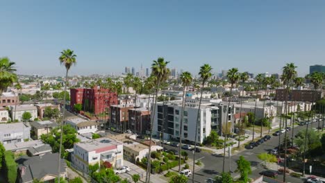 Flying-Over-a-Palm-Tree-Lined-Los-Angeles-Street-on-a-Sunny-Day,-Drone-Shot-Overlooking-Rooftops-with-Los-Angeles-Skyline-on-Horizon