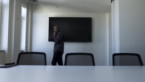 Bald-UK-Indian-Male-Talking-On-Phone-Walking-Back-And-Forth-In-Modern-Empty-Office-Meeting-Room-With-Large-Flat-Screen-TV-On-Wall
