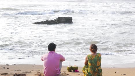 Two-people-sit-on-sandy-beach,-look-at-sea,-waves-curl-and-break-down