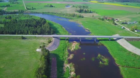 Aerial-view-of-a-Venta-river-on-a-sunny-summer-day,-lush-green-trees-and-meadows,-beautiful-rural-landscape,-wide-angle-drone-shot-moving-forward-over-the-white-concrete-bridge