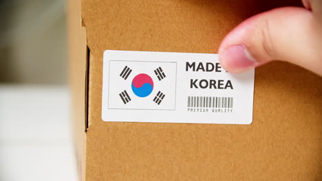 Manos-Aplicando-La-Etiqueta-De-La-Bandera-De-Corea-En-Una-Caja-De-Cartón-De-Envío-Con-Productos
