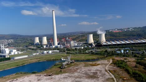 Thermal-power-plant-with-smoking-chimney,-lake-in-front-with-an-old-excavation-machine,-bright-and-sunny-afternoon