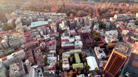 Aerial-dolly-in-of-Lastarria-neighborhood-buildings-and-Forestal-Park-autumnal-trees-in-background-at-golden-hour,-Santiago,-Chile