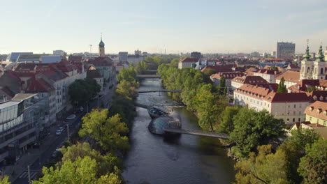 Scenic-Graz-city-Austria-aerial-view-following-Mur-river-passing-landmark-Murinsel-artificial-floating-island