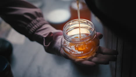 Hand-Holding-a-Glass-Jar,-Pouring-and-Fill-Fresh-Golden-Extracted-Liquid-Organic-Honey-into-it,-Traditional-Honey-Production,-Apiculture-and-Beekeeping-in-a-Bee-Farm