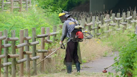 Men-workers-in-special-clothing-with-a-gasoline-mower-mow-wild-grass-near-a-wooden-fence-with-a-trimmer-in-a-residential-district-of-Seoul---slow-motion