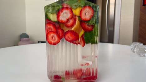 Push-In-on-Glass-Pitcher-of-Spa-Water-on-Tabletop,-Water-Infused-with-Strawberries,-Lemon,-and-Basil
