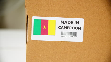 Manos-Aplicando-La-Etiqueta-De-La-Bandera-De-Camerún-En-Una-Caja-De-Cartón-De-Envío-Con-Productos