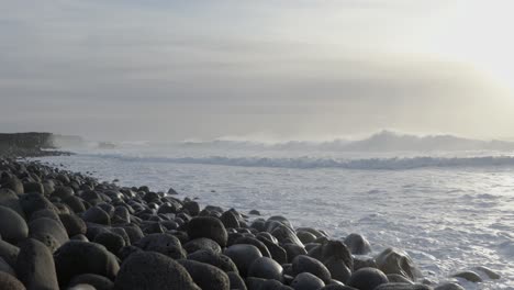 Hazy-ocean-shore-view-in-Iceland-with-waves-spraying-mist,-bright-sunshine,-rounded-stones-on-beach