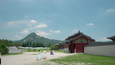 People-enter-through-Yongseongmun-Gate-to-Gyeongbokgung-Palace,-girls-in-Hanbok-dresses-walk-towards-the-entrance-with-Bukhansan-mount-summit-in-background-on-a-cloudy-day