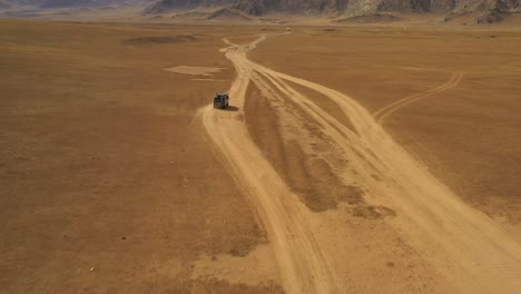 Aerial-dolly-of-van-driving-through-steppe-on-dust-road-towards-mountains-in-daytime,-Mongolia