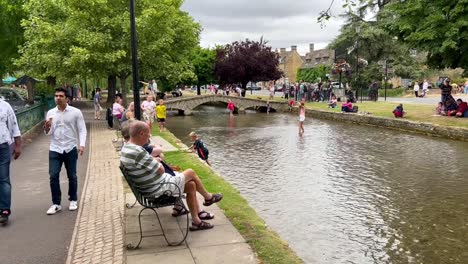 Old-couple-enjoy-sitting-by-the-riverside-where-the-river-Windrush-passes-though-the-village-centre-in-Bourton-On-The-Water,-Cotswolds---England,-UK