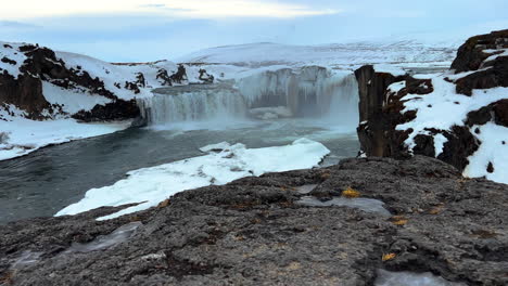 Slow-motion-Tilt-up-shot-of-Goðafoss-Waterfall-surrounded-by-snowy-hills-on-Iceland-during-daytime