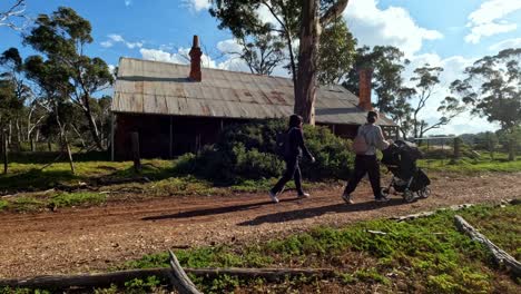 Woman-walking-along-a-dirt-track,-pushing-a-stroller-past-an-old-abandoned-heritage-tin-roof-house-in-regional-Victoria