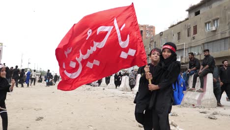 Two-kids,-a-boy-and-a-girl,-are-raising-red-flags-during-the-Muharram-juloos-or-parade,-also-known-as-Muharram-Ul-Haram,-which-is-considered-to-be-the-second-holiest-month-after-Ramadan