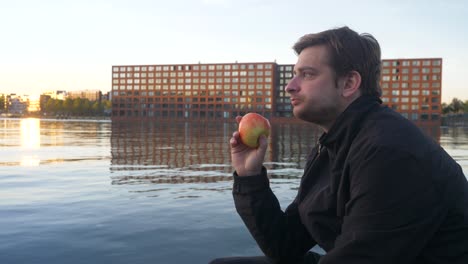 Junger,-Gutaussehender-Mann-In-Einer-Schwarzen-Jacke,-Der-Bei-Sonnenuntergang-Einen-Roten-Apfel-Auf-Einem-Städtischen-Pier-In-Amsterdam-Isst