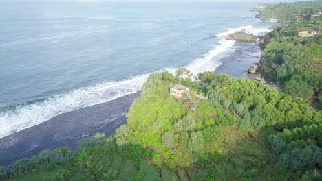 Orbit-drone-shot-of-waves-reaching-green-coastline-of-Indonesia-with-private-house-on-the-top-of-cliff
