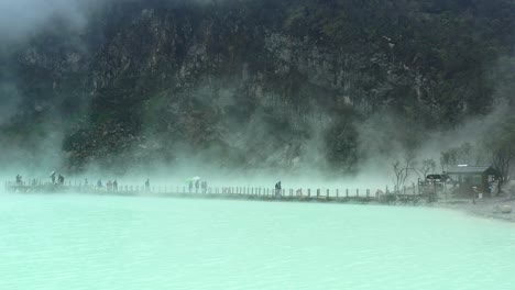people-walking-on-a-bridge-with-steam-rising-from-sulfur-lake-and-mountain-behind,-aerial
