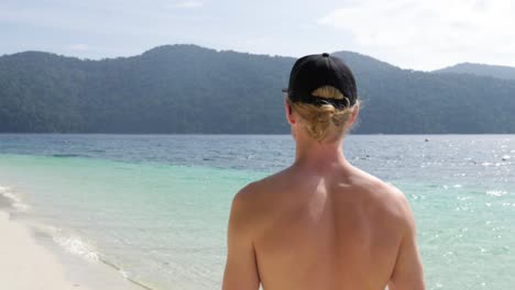 Ultra-Zeitlupenaufnahme-Eines-Jungen-Kaukasischen-Mannes-Mit-Mütze,-Der-Am-Atemberaubenden-Strand-In-Thailand-Spaziert-Und-Auf-Das-Meer-Blickt