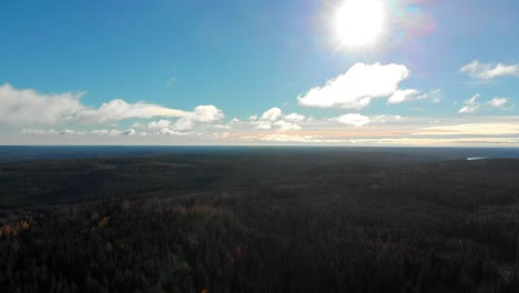 Drone-footage-flying-over-a-colourful-autumn-forest-towards-the-bright-sun,-a-blue-sky-and-clouds