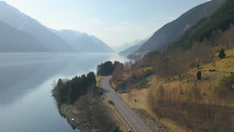 Luftaufnahme-Einer-Leeren-Straße,-Die-Sich-Am-Ufer-Eines-Fjords-Entlang-Schlängelt,-Mit-Wunderschönen-Bergen-Im-Hintergrund-In-Norwegen