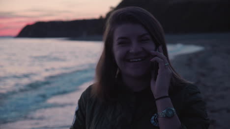 Girl-receiving-a-phone-call-and-talking-on-the-phone-whie-walking-along-the-beach-on-the-sunset-with-sea-waves-crushing-on-shore-in-the-background