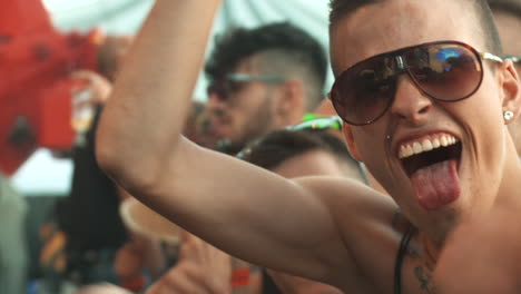 Spanish-looking-male-with-chest-tattoos-and-tinted-sunglasses-cheers-at-a-music-festival-with-his-tongue-out-and-waves-his-arms
