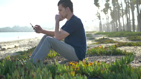 A-young-white-man-sits-on-the-beach-looking-at-his-smart-phone-while-seeming-nervous,-upset,-or-stressed-out-in-Santa-Barbara,-California