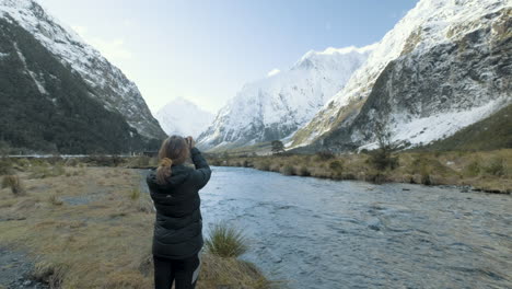 Girl-photographing-river-surrounded-by-snow-capped-mountains-in-New-Zealand