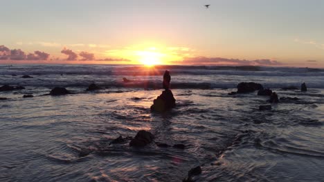 Atemberaubender-Blick-Auf-Den-Sonnenuntergang-Bei-Ebbe-Am-Laguna-Beach-Mit-Einer-Fliegenden-Drohne-Und-Surfenden-Surfern-Im-Hintergrund,-Während-Die-Sonne-Zwischen-Den-Felsen-Ihren-Höhepunkt-Erreicht-Und-Vom-Wasser-Reflektiert-Wird