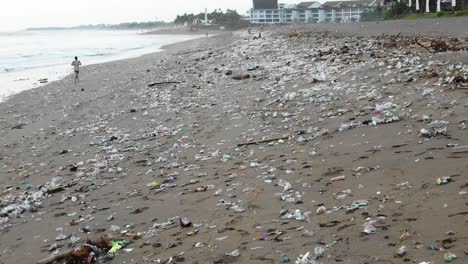 Tracking-shot-of-a-man-jogging-on-a-badly-polluted-beach,-tons-of-plastic-garbage-on-the-shore