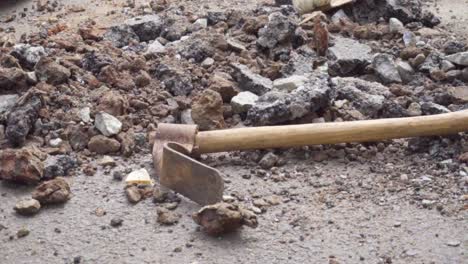rusty-old-digging-tool-shaped-like-a-pickaxe-with-a-forged-adze-isolated-against-a-rock-and-tar-debris,-slow-motion-pan