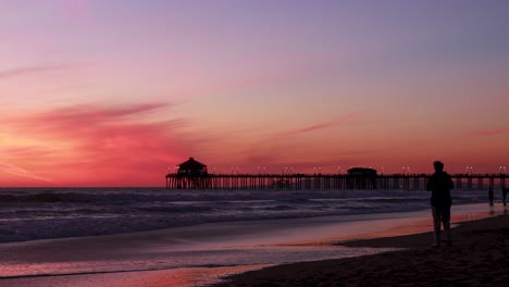 A-man-enjoys-his-vacation-at-the-beach-during-a-gorgeous-red,-purple,-tangerine,-pink-and-blue-sunset-with-the-Huntington-Beach-Pier-in-the-background-at-Surf-City-USA-California