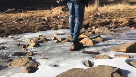 Man-wearing-boots-stepping-over-rocks-on-frozen-river